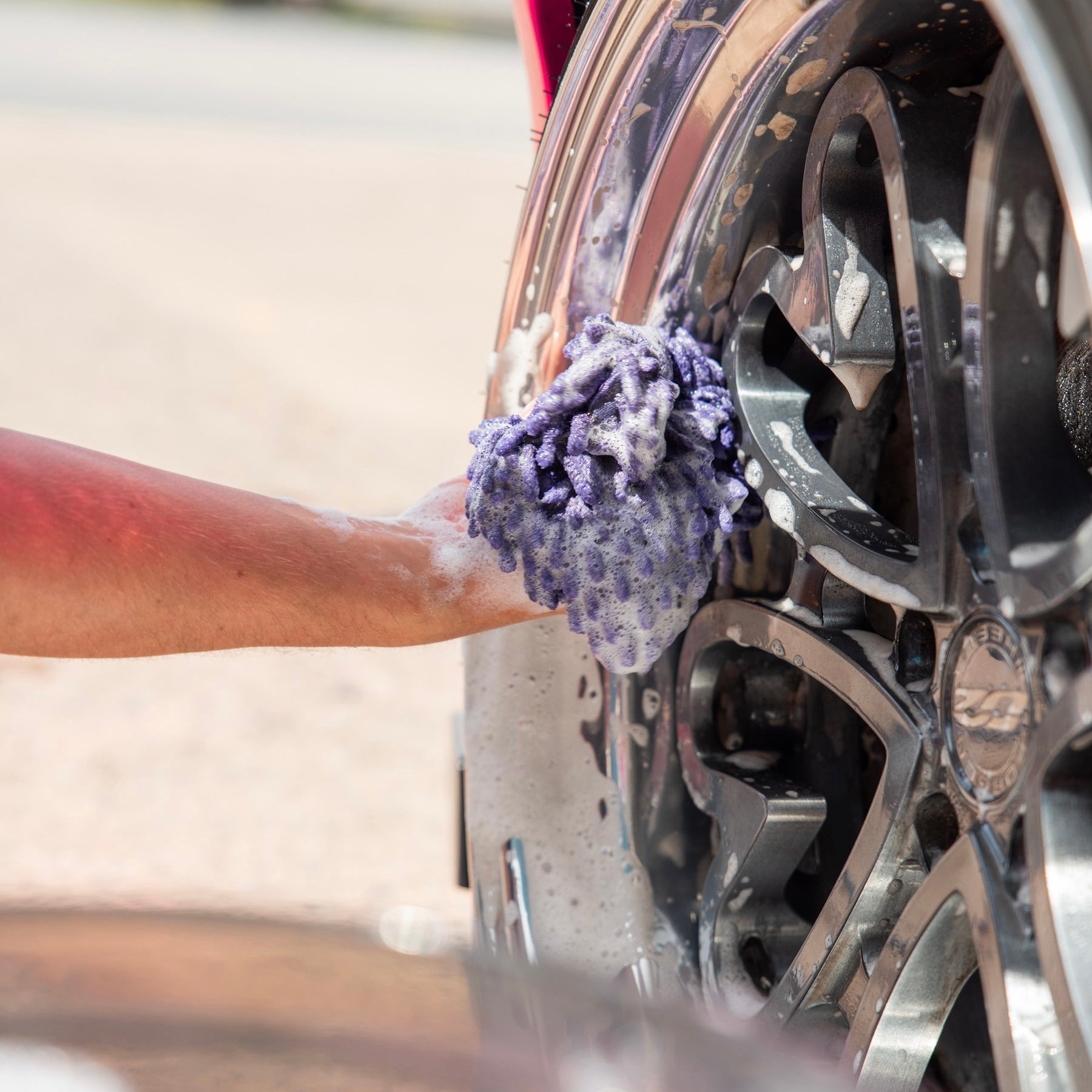 Person cleaning a car wheel with a purple sponge outdoors using wheel shampoo