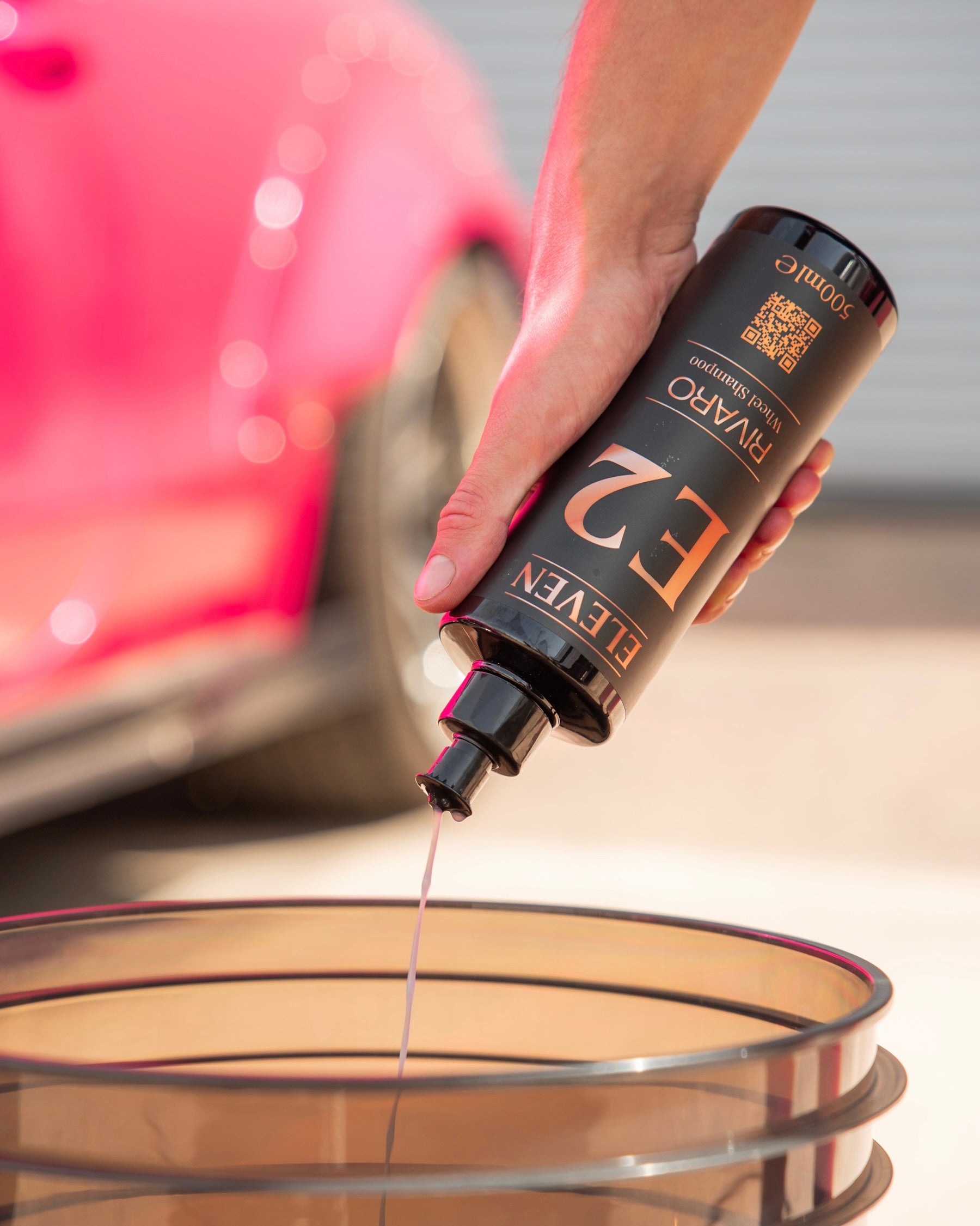 Hand pouring a wheel shampoo from a black bottle labeled 'E2' into a wash bucket with a pink car in the background.