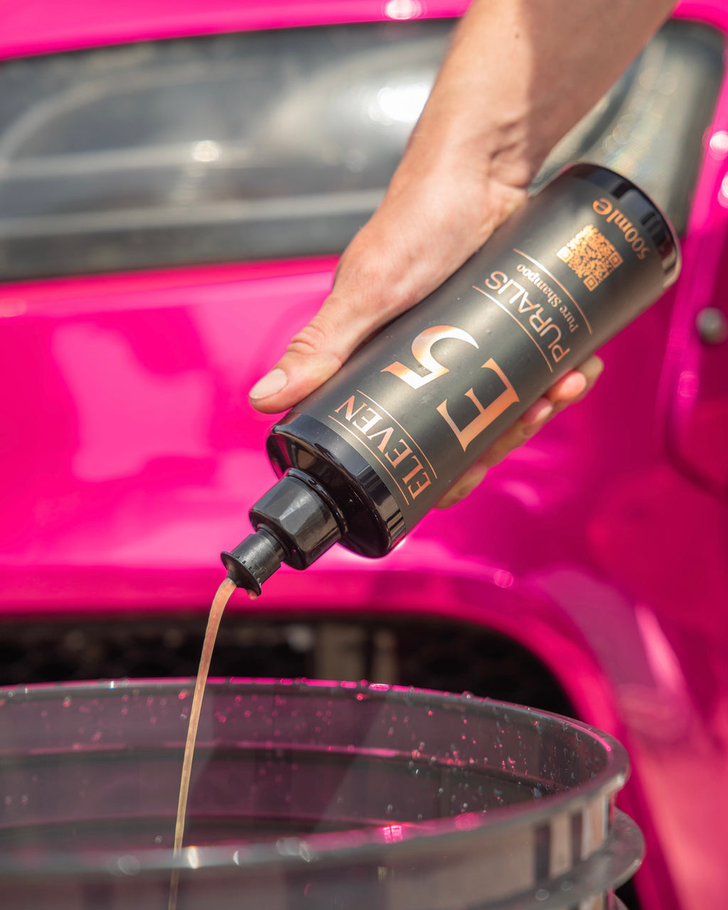 Person pouring E5 liquid from an ELEVEN bottle into a black bucket with a pink car behind