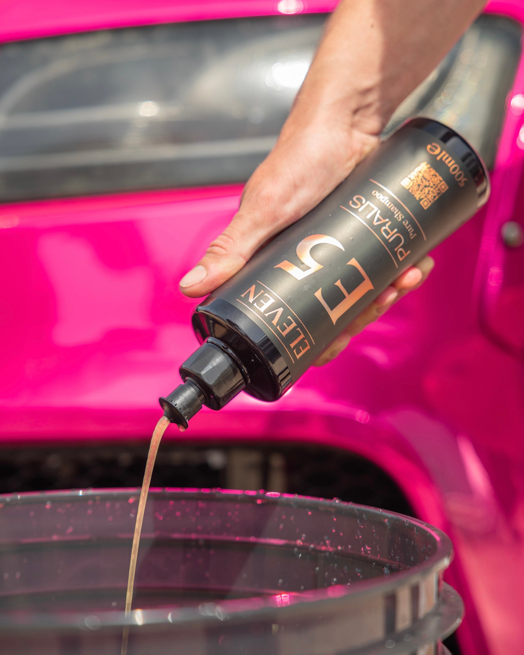 Person pouring E5 liquid from an ELEVEN bottle into a black bucket with a pink car behind