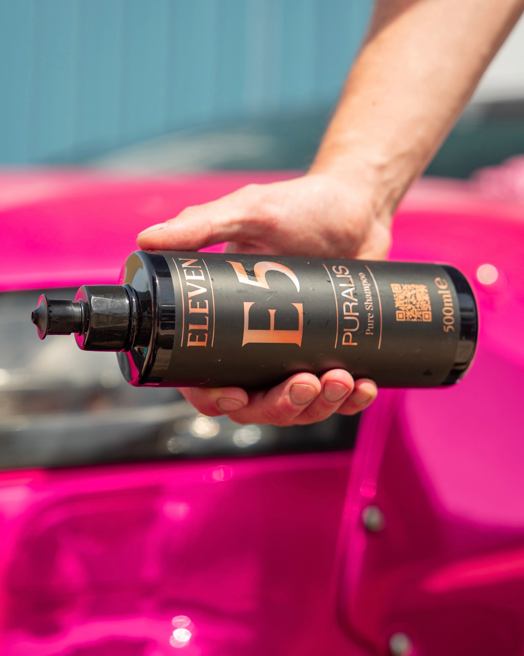 Hand holding an ELEVEN E5 PURALIS bottle in front of a pink car panel