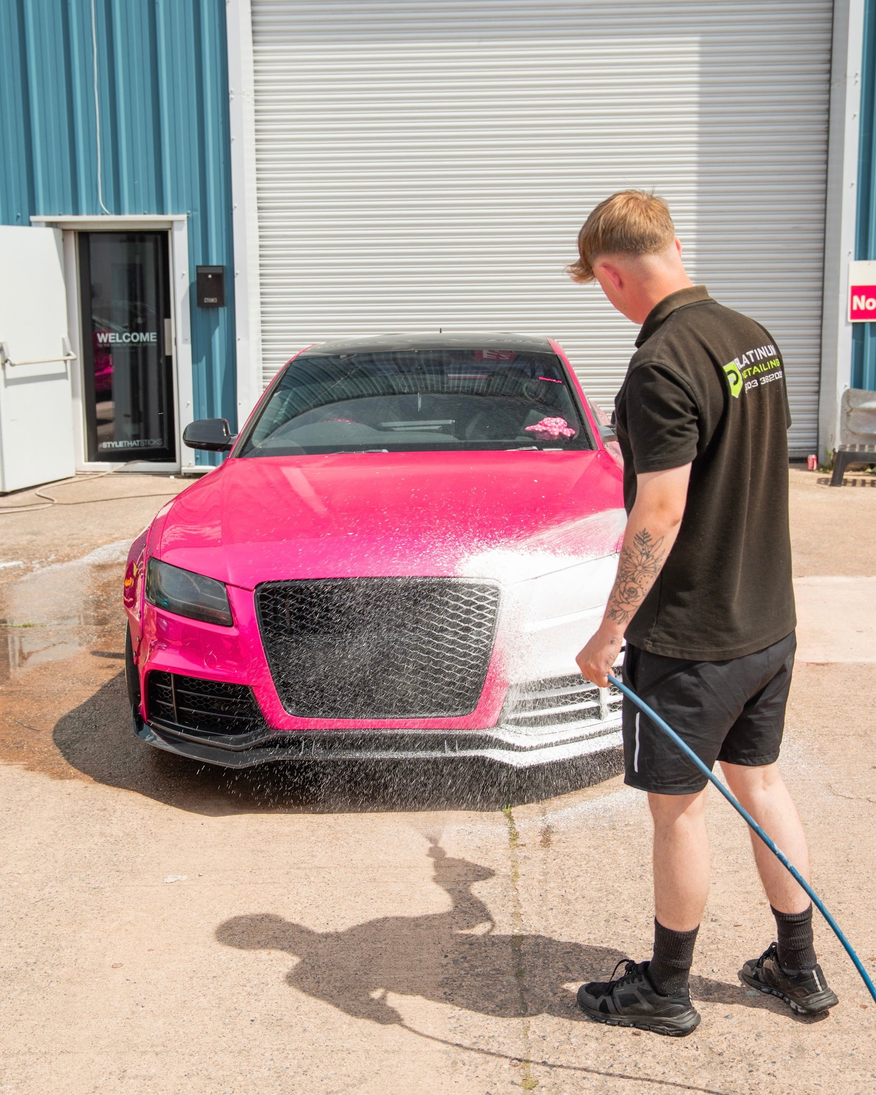 Person washing a pink car with a hose outside a detailing workshop