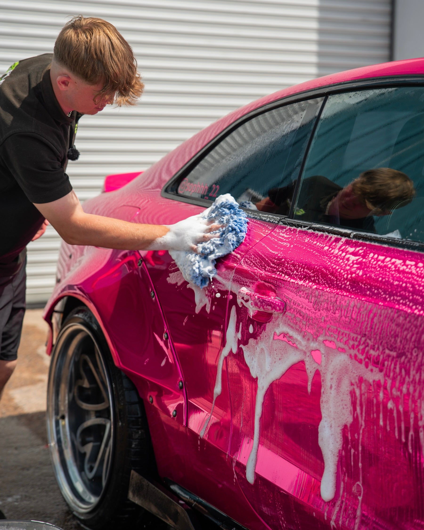 Person washing a pink car with a blue sponge and soapy water near the rear wheel