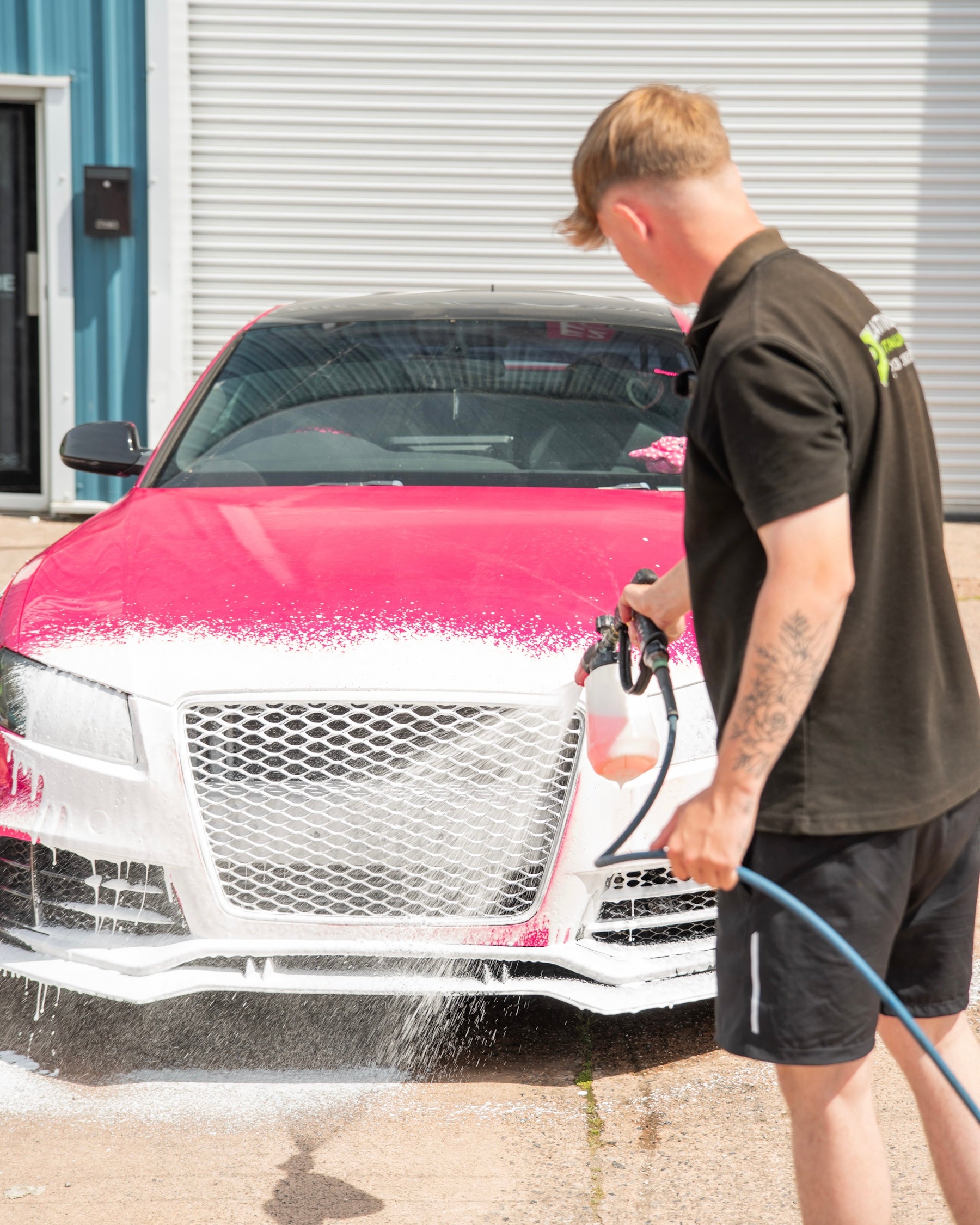 Person washing a pink car with a pressure washer in front of a building.